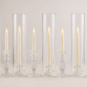 Elegant wedding table featuring clear glass hurricane sleeves, ribbed glass candle holders, flameless taper candles, white roses, and greenery arranged as a romantic centerpiece display.