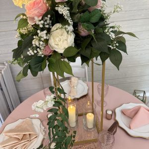 Tall gold centerpiece with pink, ivory, and white artificial flowers, greenery, and two cylinder vases with flameless candles