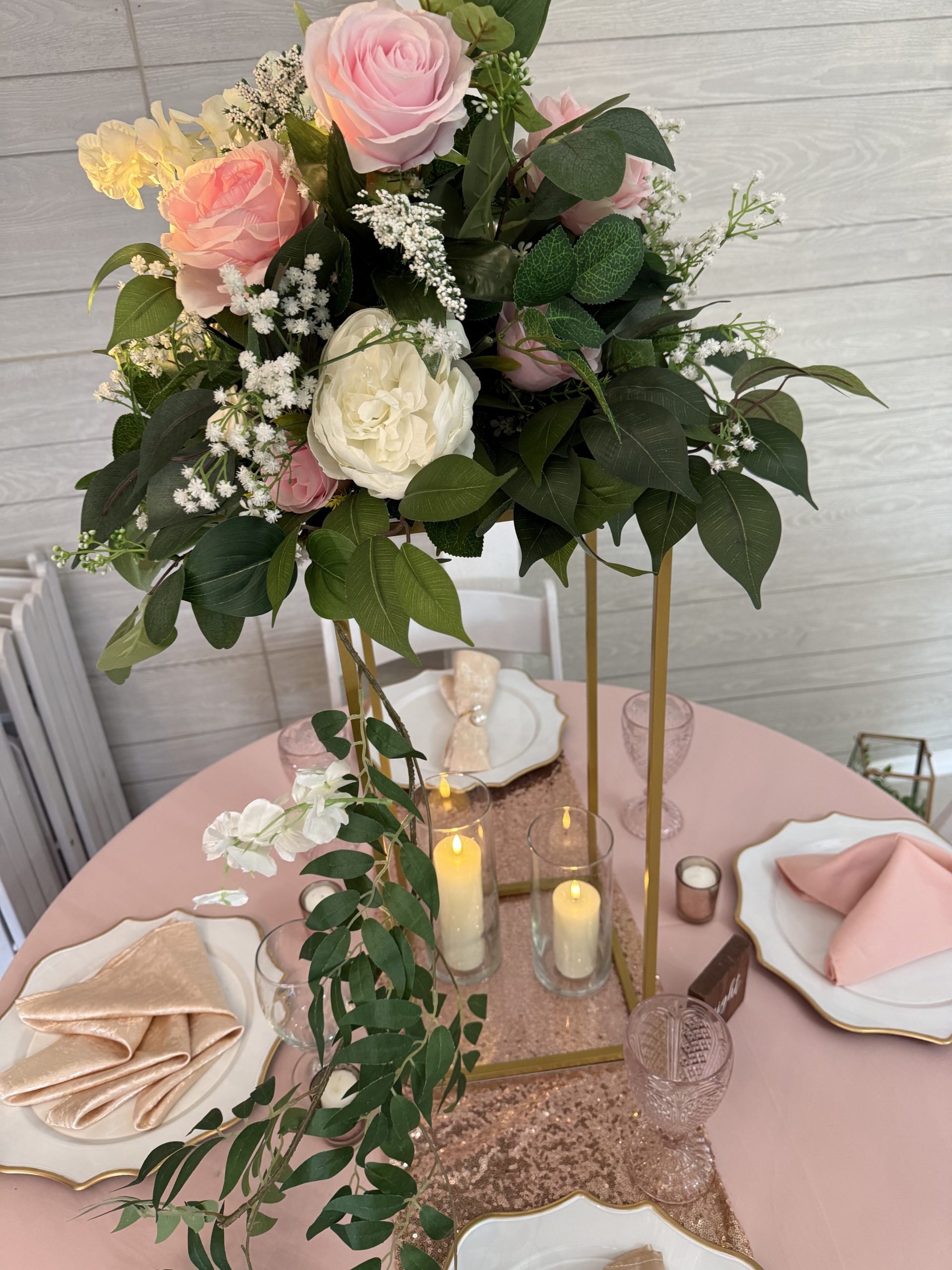 Tall gold centerpiece with pink, ivory, and white artificial flowers, greenery, and two cylinder vases with flameless candles