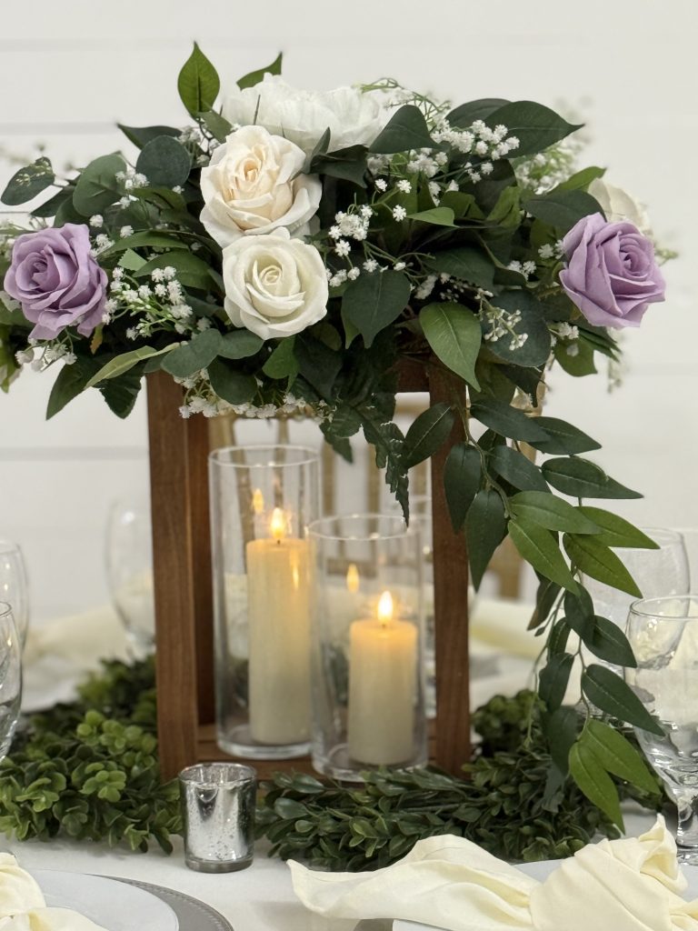 Rustic wooden lantern centerpiece rental with custom artificial floral arrangement in ivory, white, and accent color, surrounded by greenery, cylinder vases with flameless candles, and three votives, available in Leander, Texas.